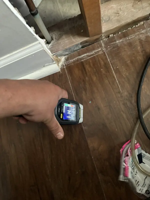 A handheld moisture meter checking flooring beside an opened wall and exposed plumbing